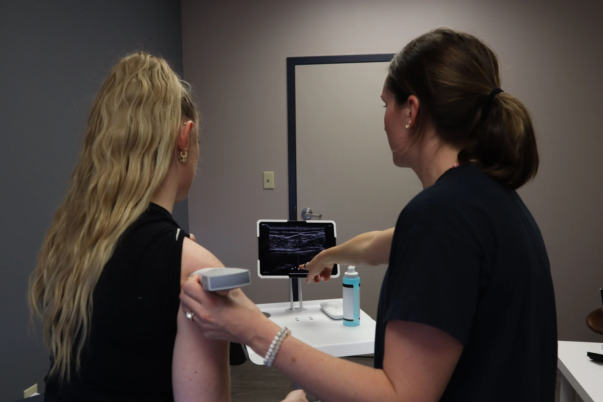 Illustrative stock photograph: a clinician scans a patient's shoulder with a handheld ultrasound probe while pointing to the tablet display.